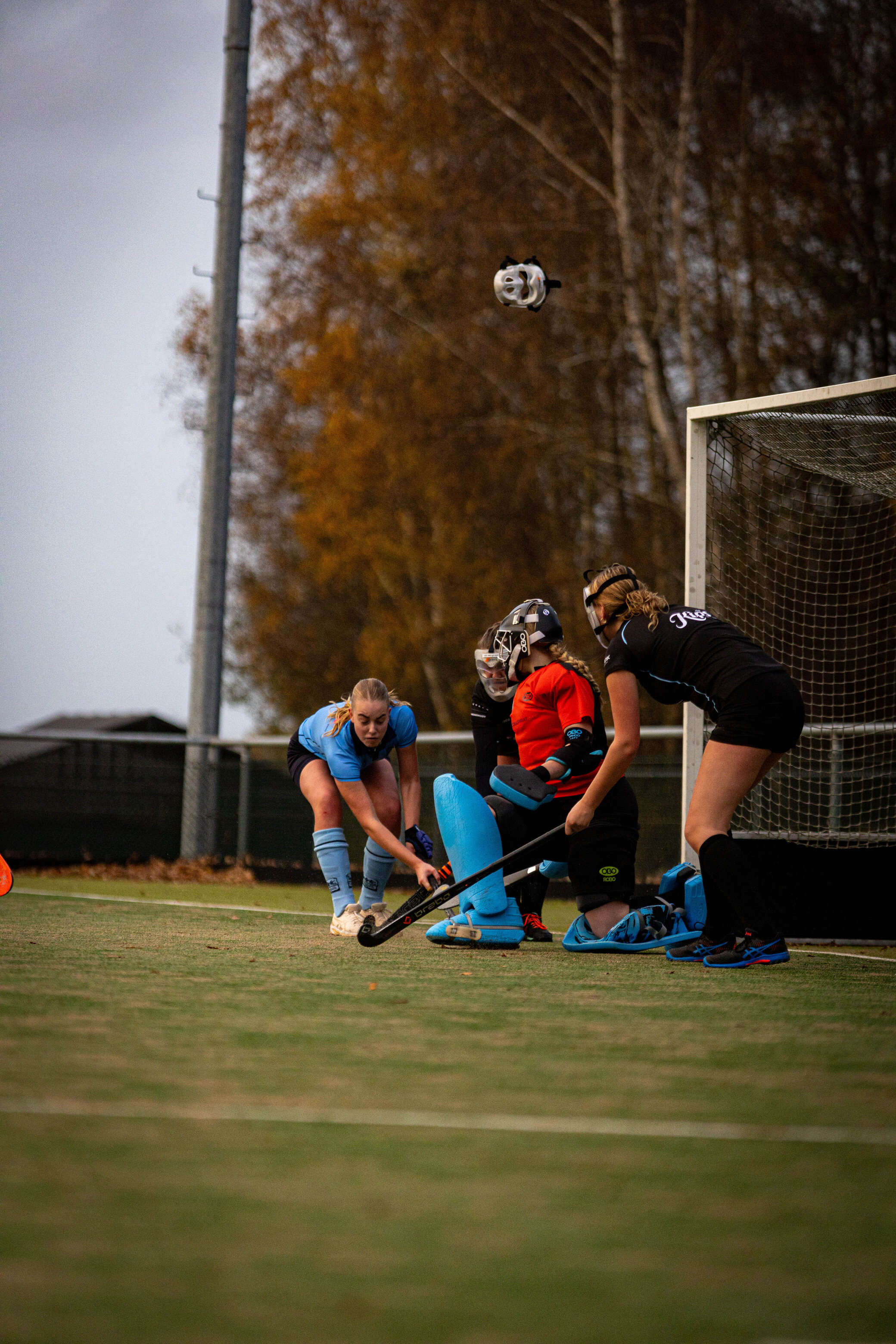 A hockey game with 3 women wearing blue and a woman in an orange shirt. They are playing near the net.