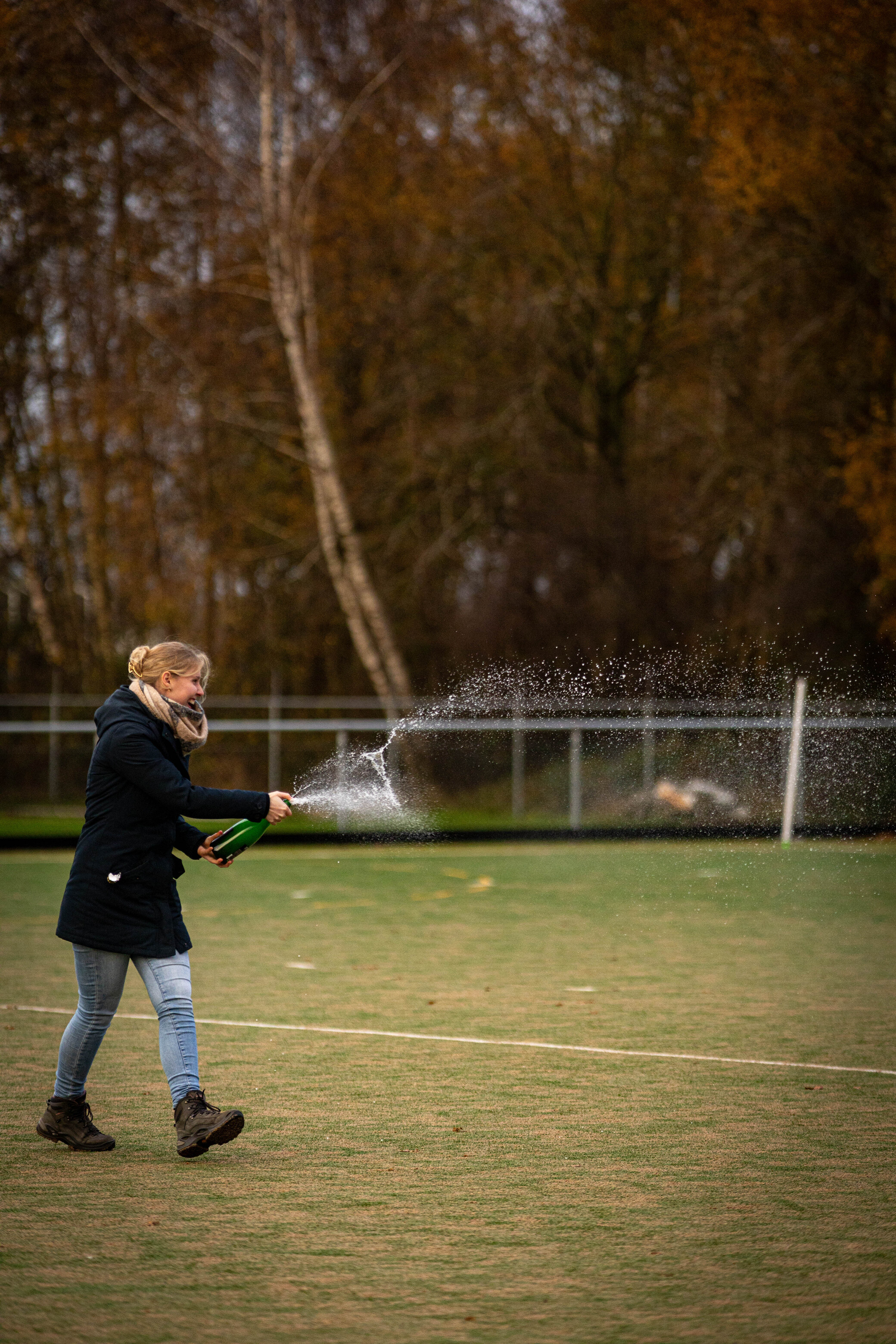 A girl is holding a bottle of champagne and spraying it in the air at an outdoor hockey rink.