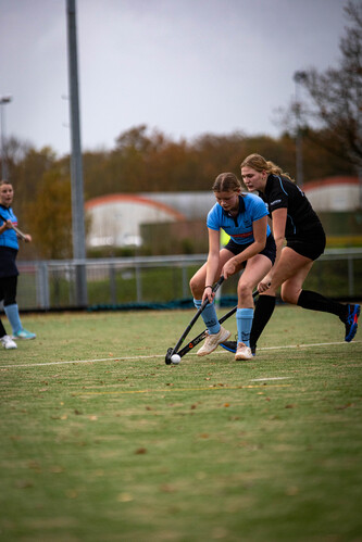 Two women playing hockey, the player on the left is wearing a blue shirt and white shorts.