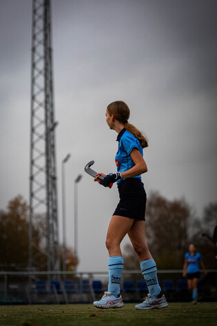 A female hockey player wearing blue and white uniform is walking on a field with her stick.