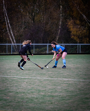 A girl in a blue uniform is about to hit the puck with her hockey stick while another girl in a black uniform blocks her way.