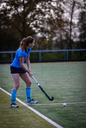 A young woman in a blue hockey uniform is getting ready to play. She's wearing knee pads and her hands are on the ice stick.