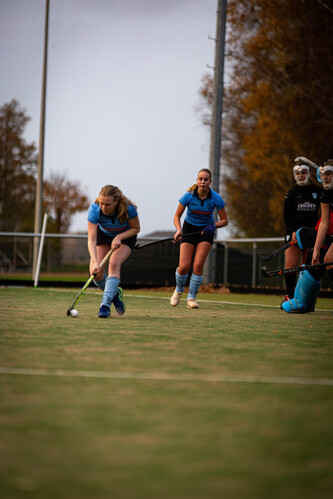 Three women playing hockey on a field in front of trees.