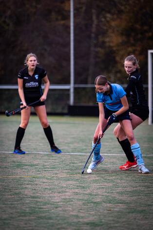 Three girls play hockey on a field with numbers 19, 16, and 18.