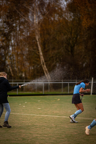 A girl in a blue jersey is spraying water on the ground, and another girl watches from behind her.