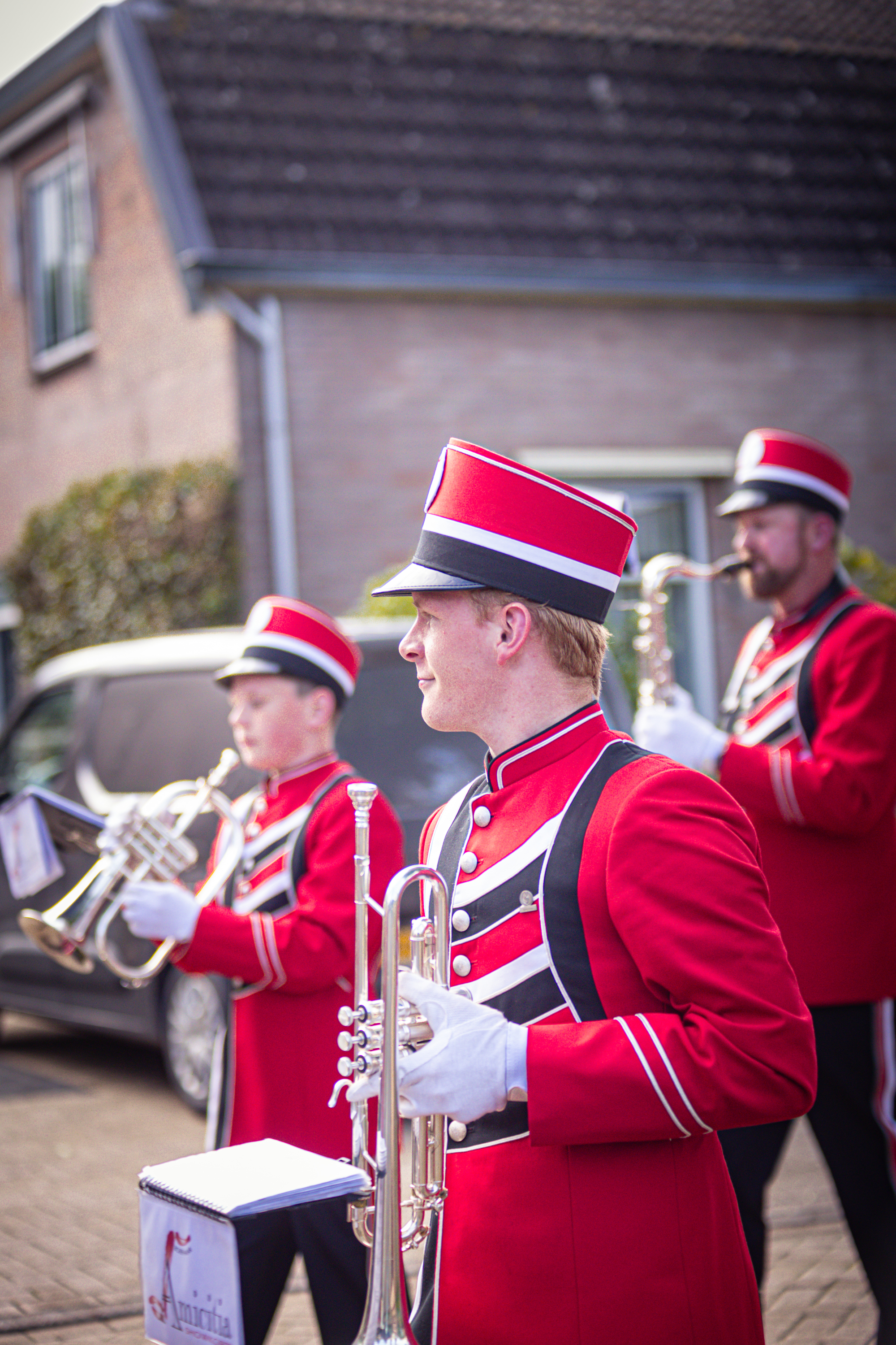 Three brass musicians in matching red and white uniforms stand in front of a building, their instruments at the ready.