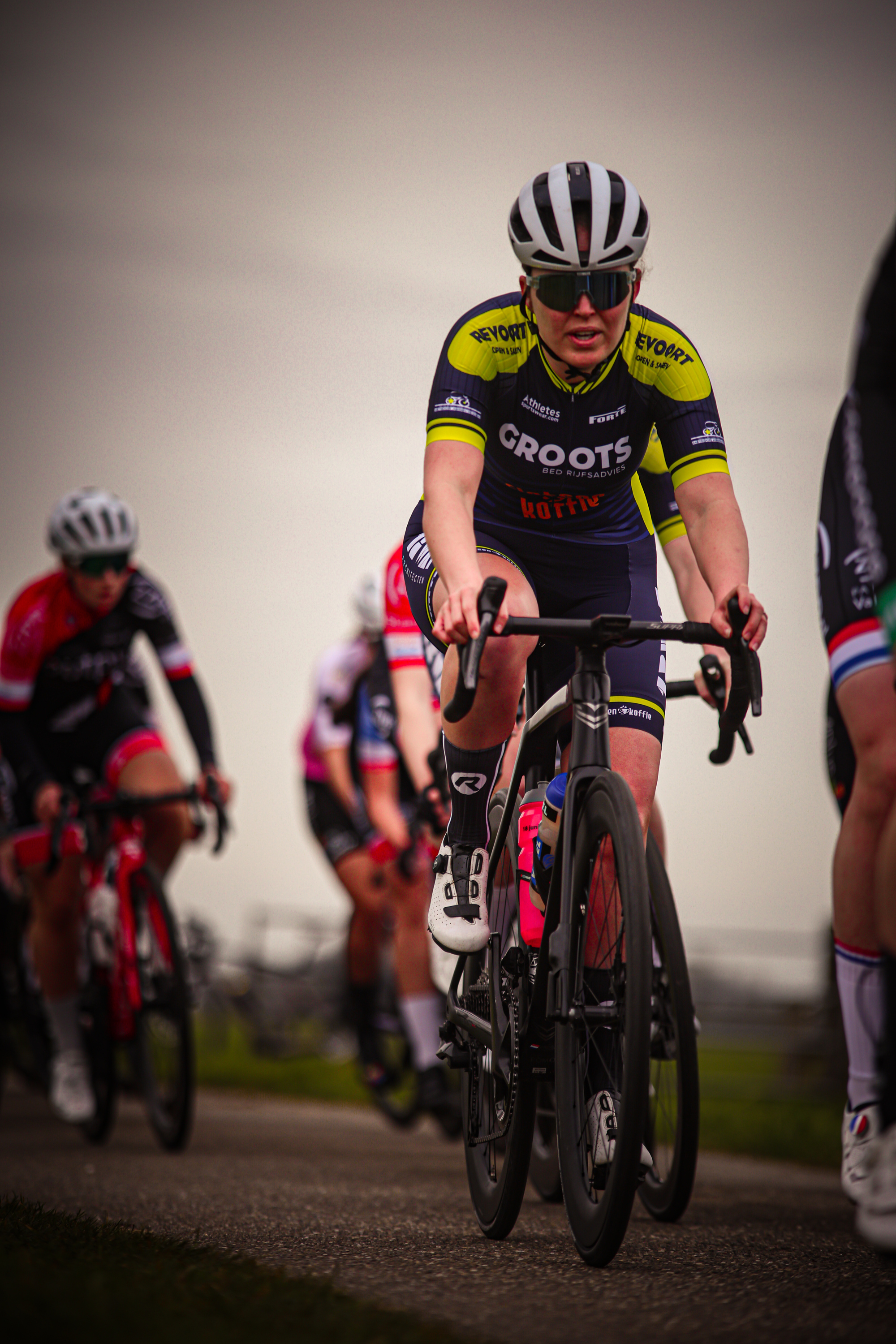 Several cyclists race in a group, with one wearing a yellow and black shirt that says "Wielrennen".