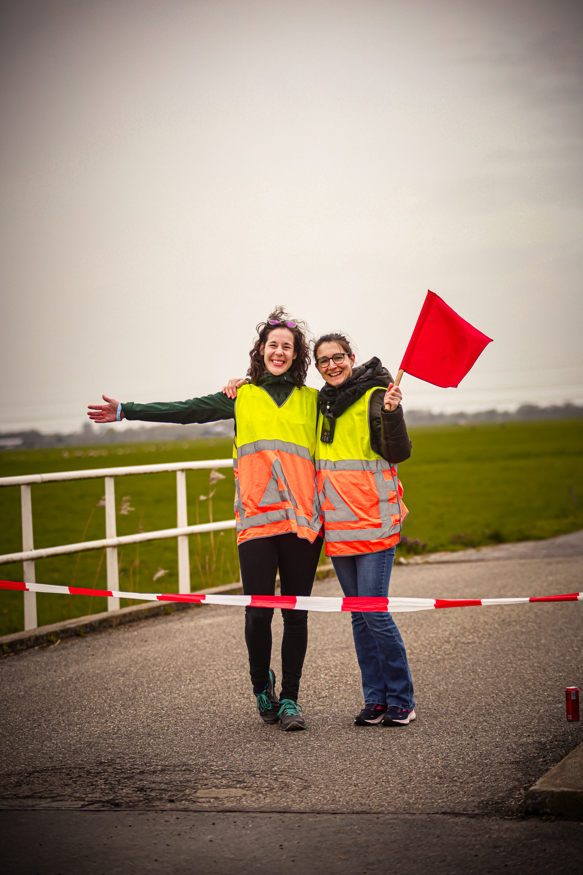 Two women are standing in a grassy field, holding red flags. They both have on safety vests and are smiling at the camera.