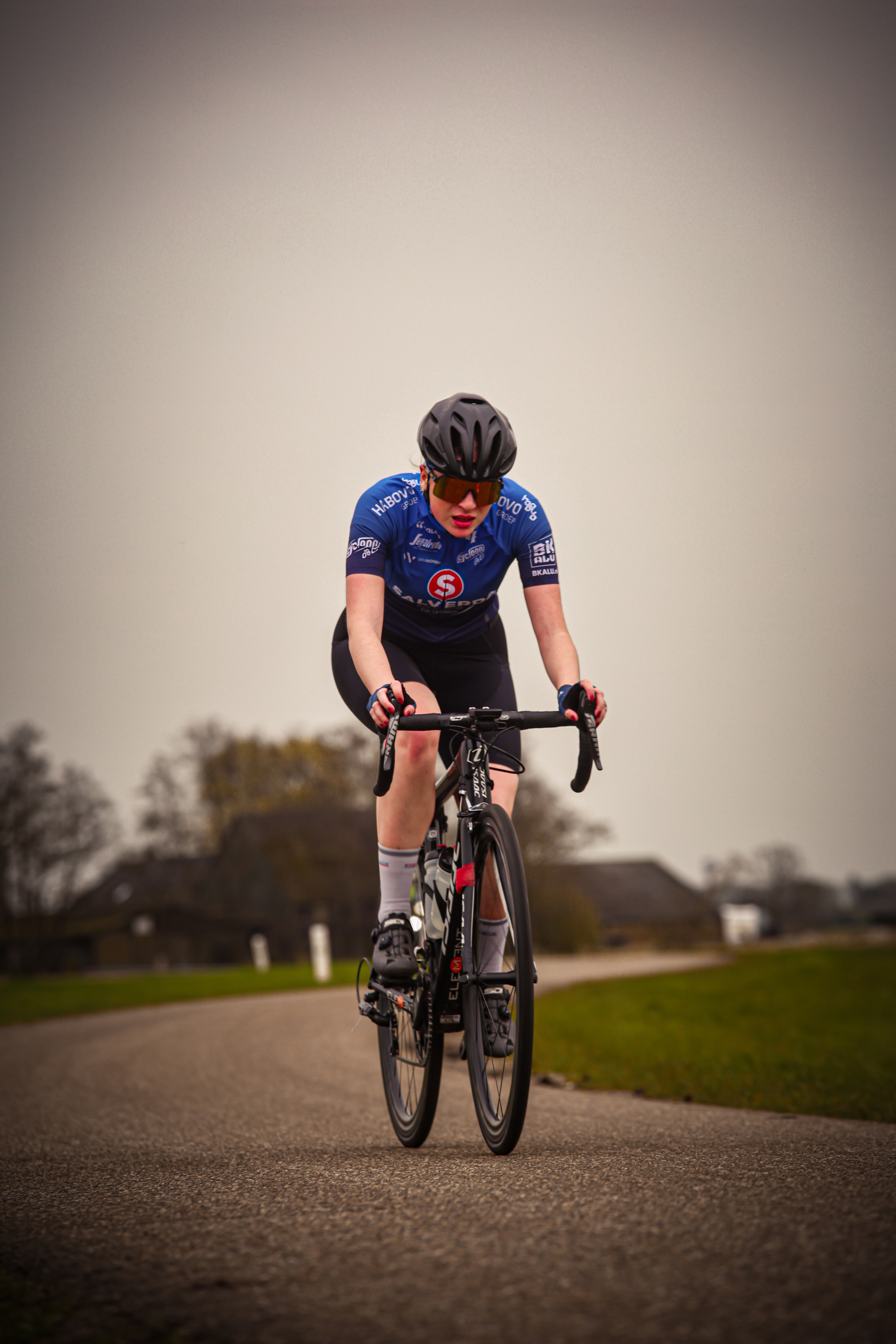 A woman on a bicycle in the middle of a road with a blue top saying "wielrennen" on it and she is wearing sunglasses.