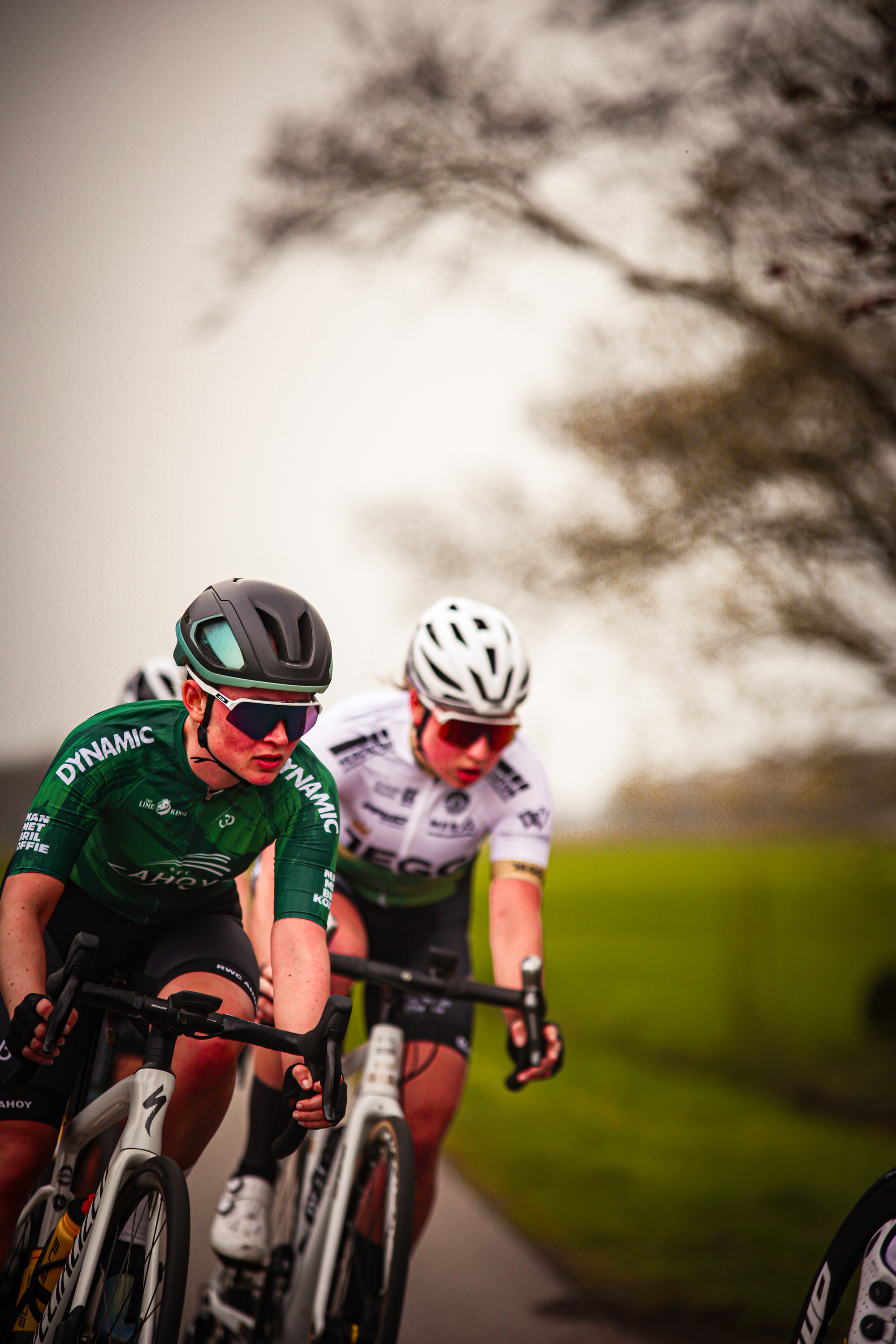 Two cyclists in green and white jerseys ride down a road with a grassy area to the right.