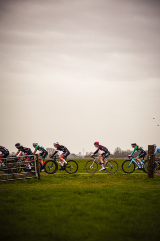 A group of cyclists wearing black helmets are racing on a grassy field during the Ronde van de Lichtmis race.