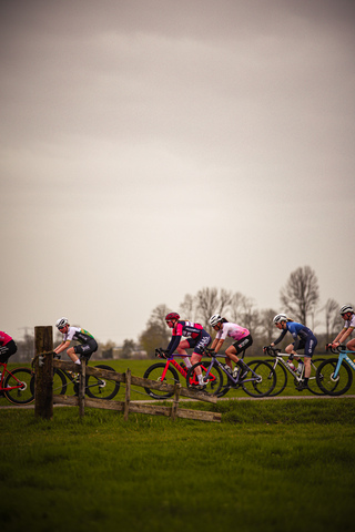 A group of cyclists are racing through a grassy field, with trees in the background. The sky is cloudy and it's raining.