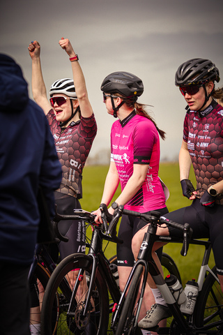 A group of cyclists are riding side by side in a grassy field with one cyclist proudly lifting their arms to celebrate.