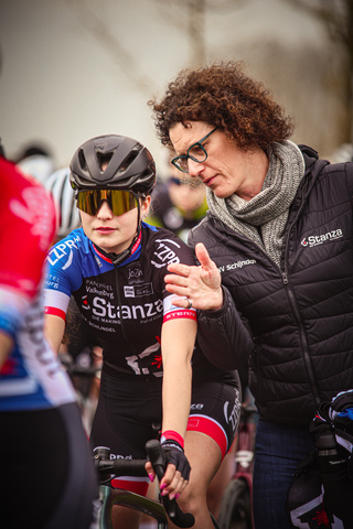 Two women on their bikes at a race, with one wearing a bib that says STANZA on it.