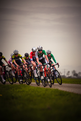 A group of 6 cyclists in motion on a grassy field, with the cyclist in the lead wearing a red jersey.