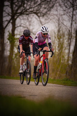 Two women riding bikes on a road together with one wearing a pink shirt.