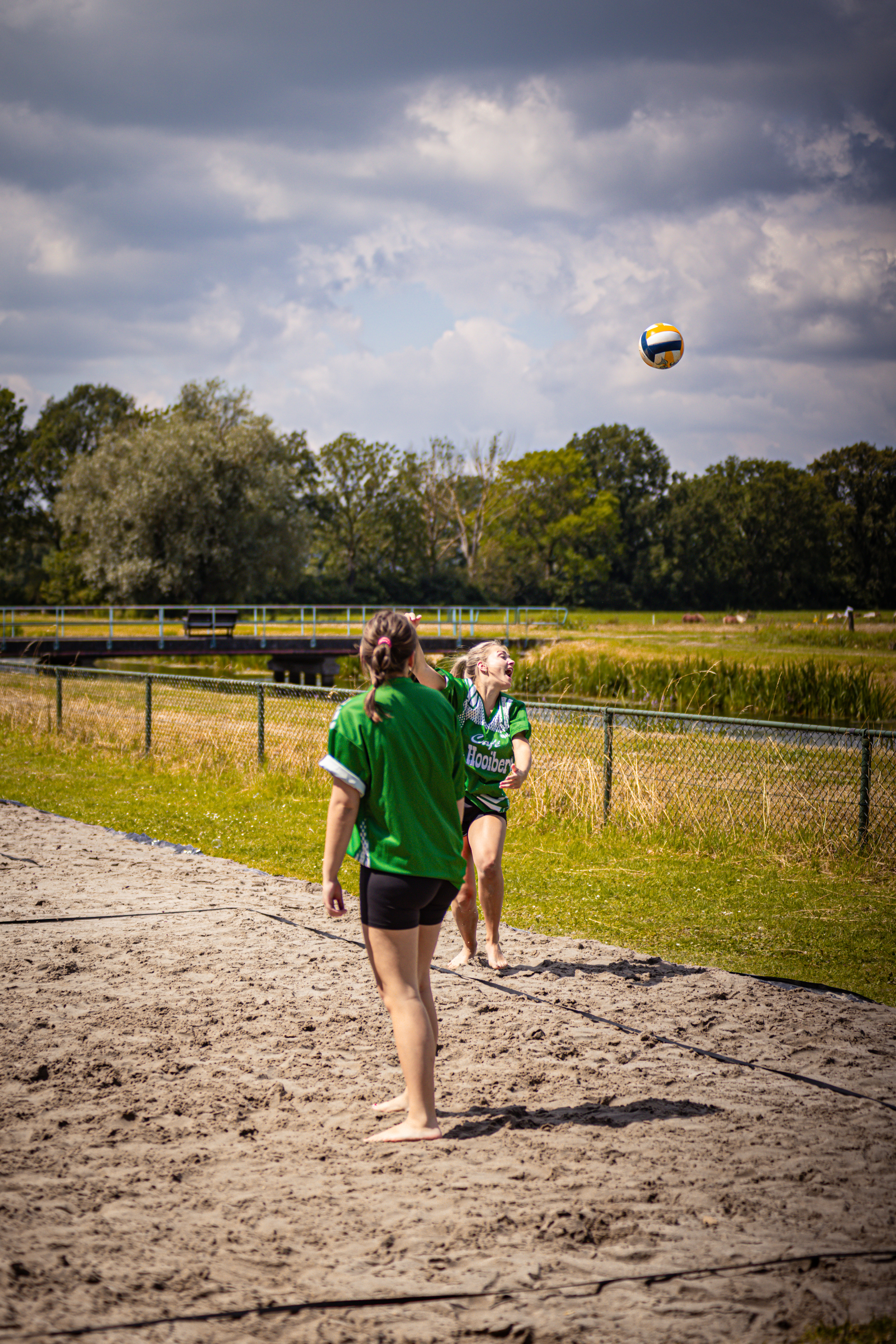 Two girls are playing a game of beach volleyball, one in green and one in blue. They're both jumping up to hit the ball.