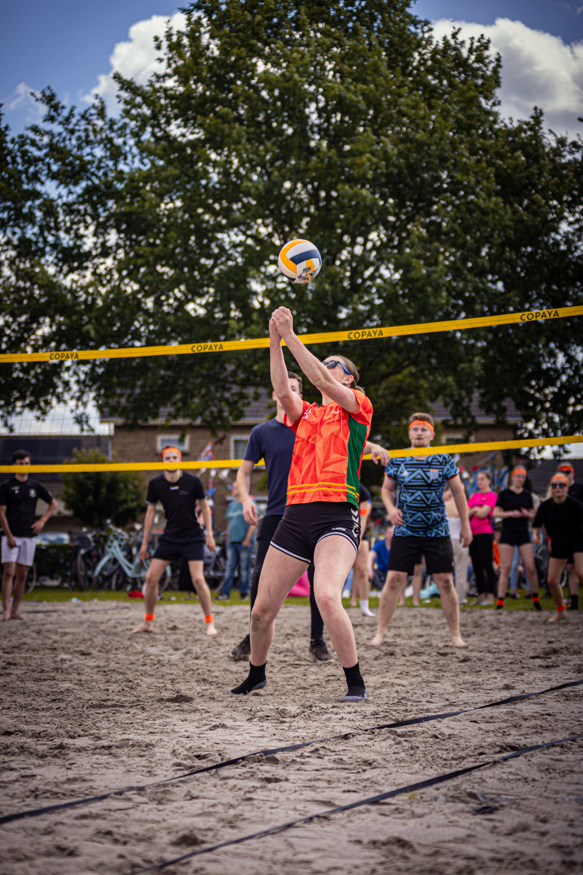 A group of people playing beach volleyball, one of the players is wearing an orange shirt.