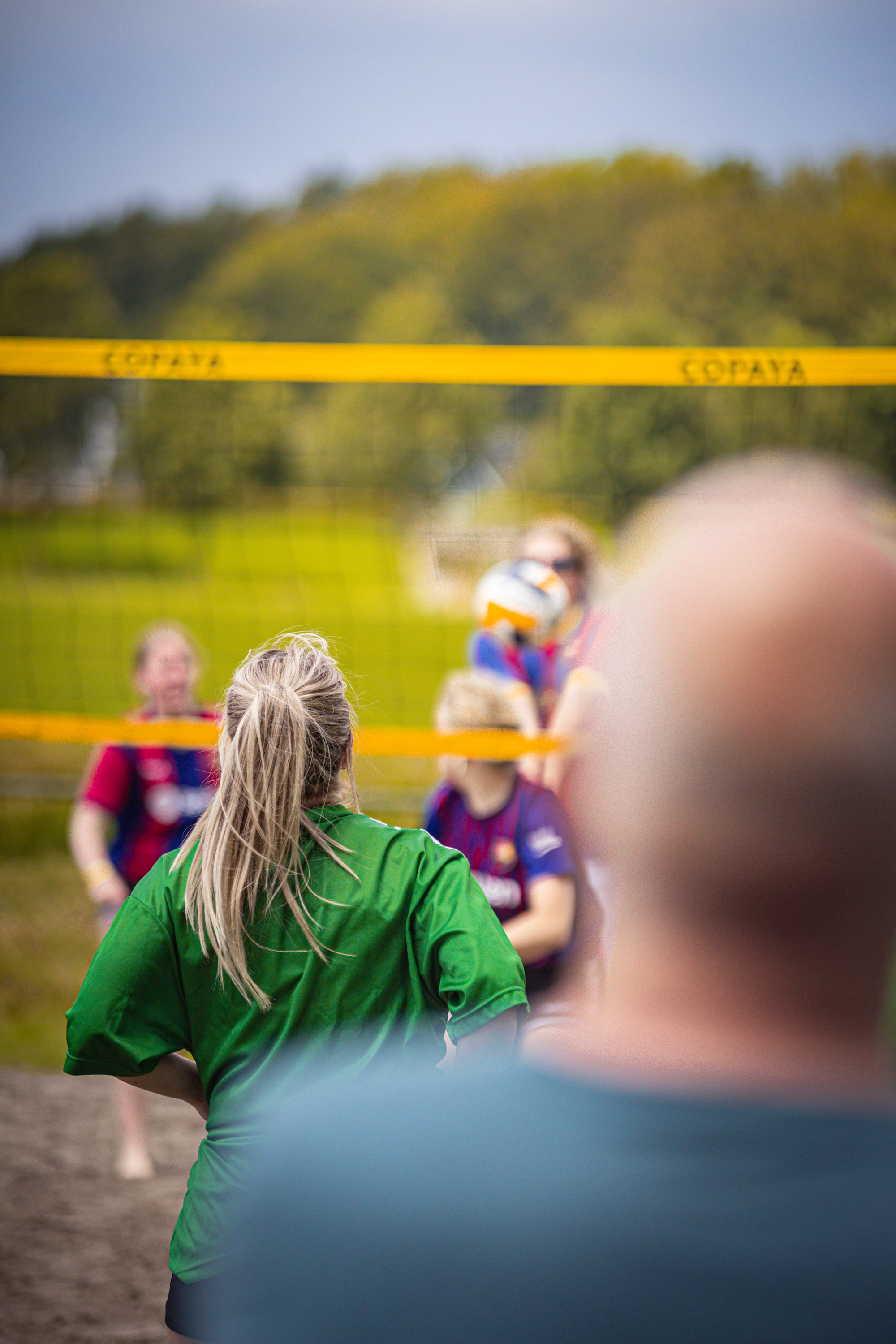 A group of people are playing volleyball on a beach, with a man wearing a blue shirt watching from the side.