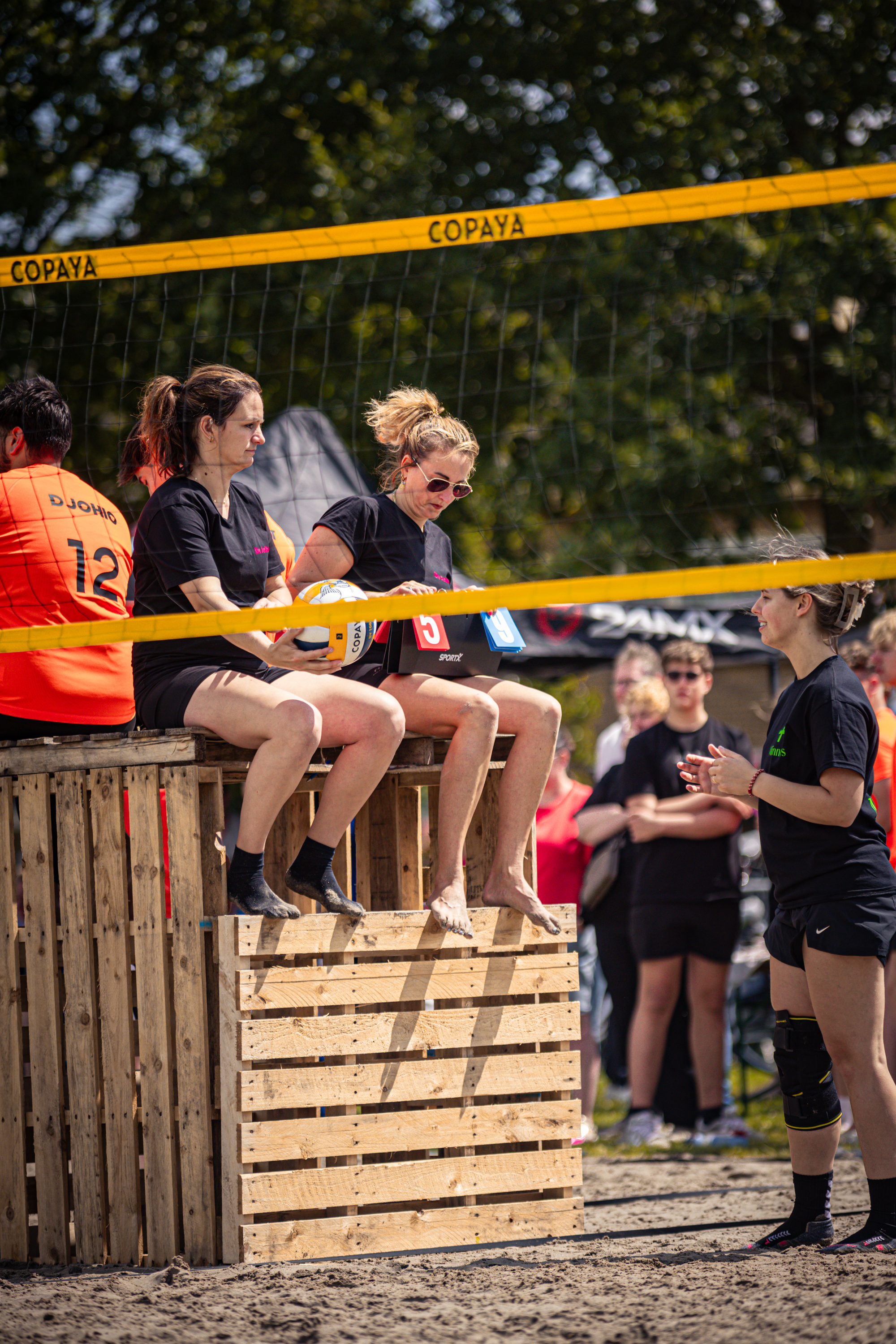 Five people sitting on a wooden box with a volleyball net in the background.