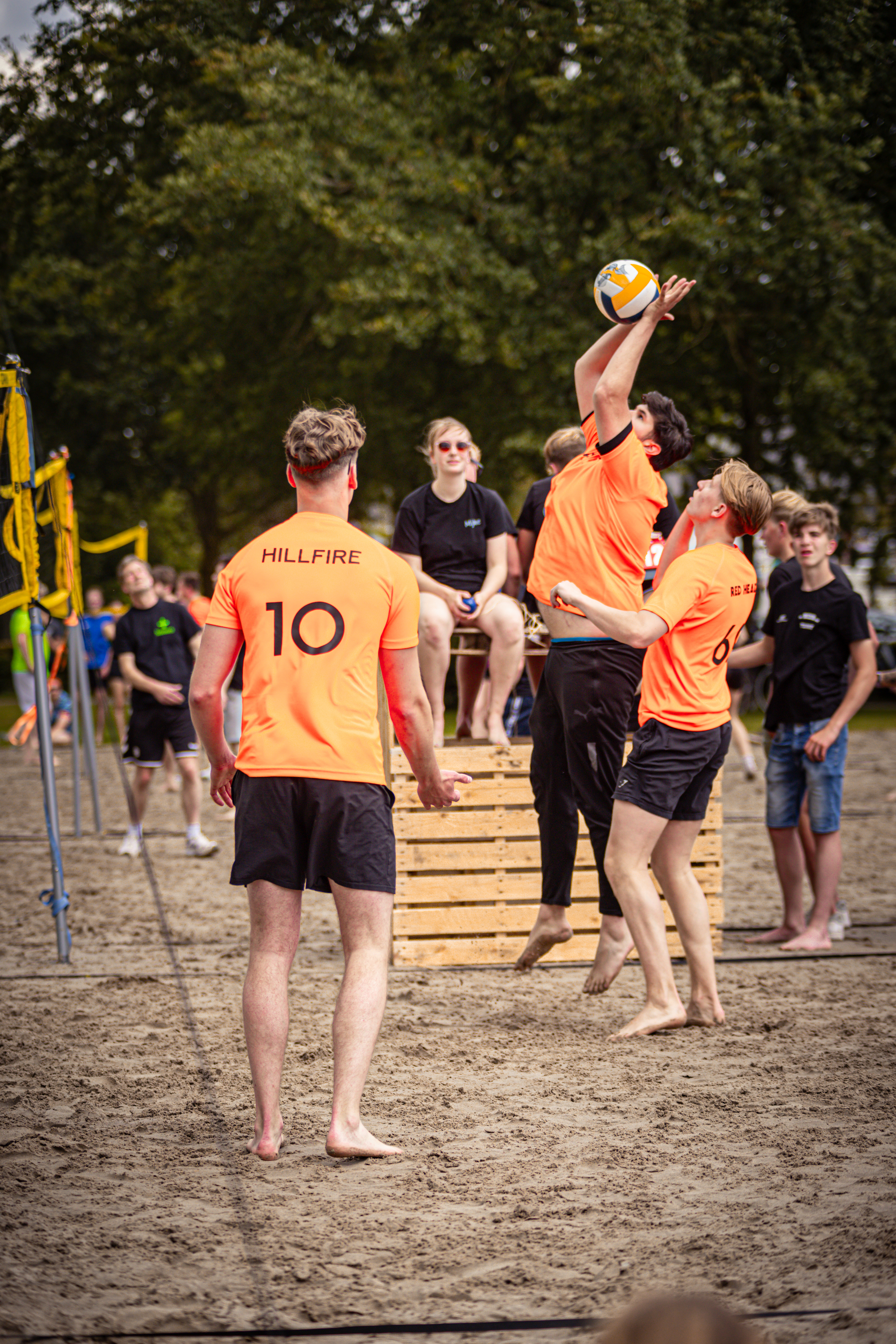 Several people are playing beach volleyball in an orange and black outfit. The ball is airborne while two men jump to hit it.