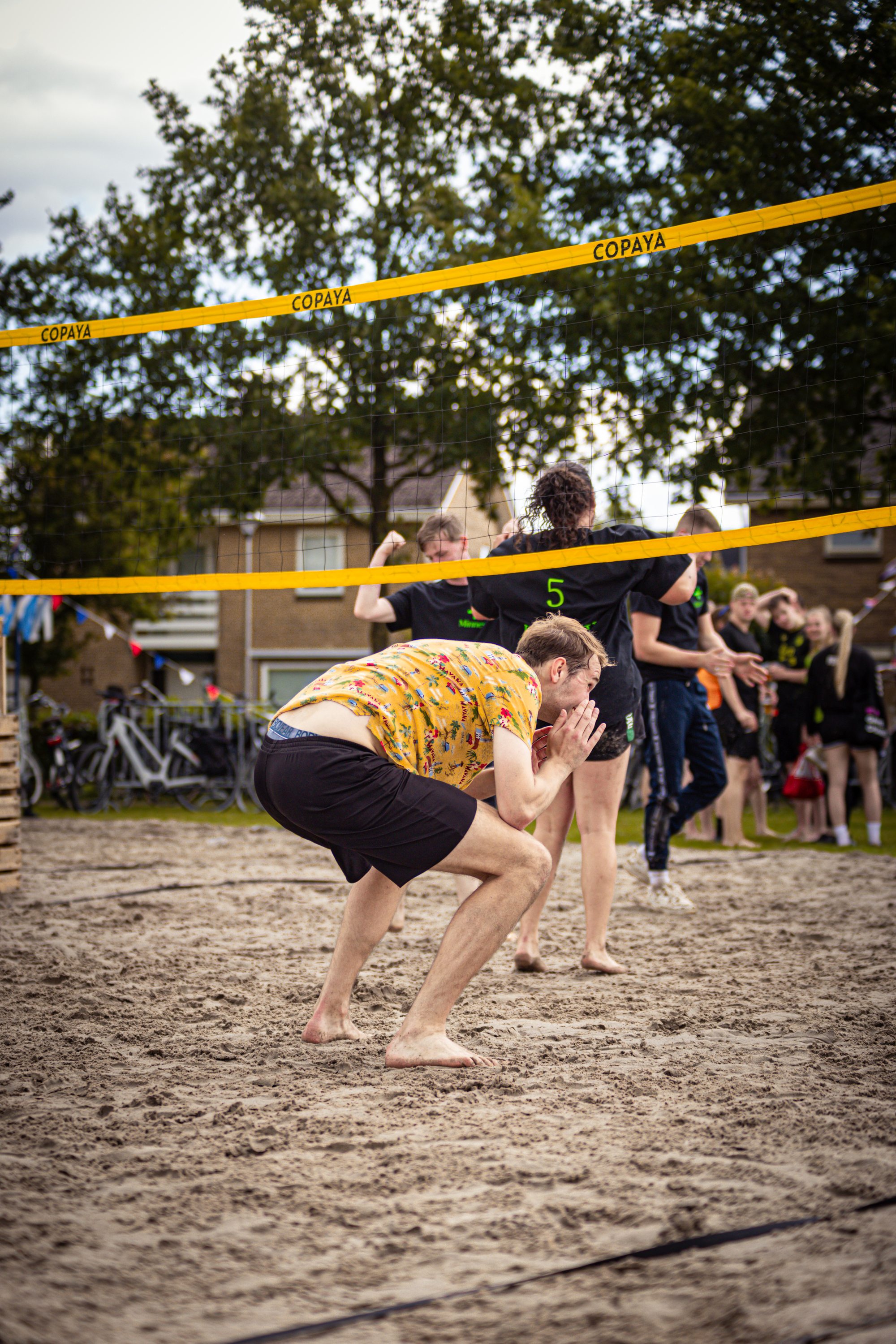 A man in a yellow shirt and black shorts kneels on a sandy beach, preparing to play volleyball with a group of other players.