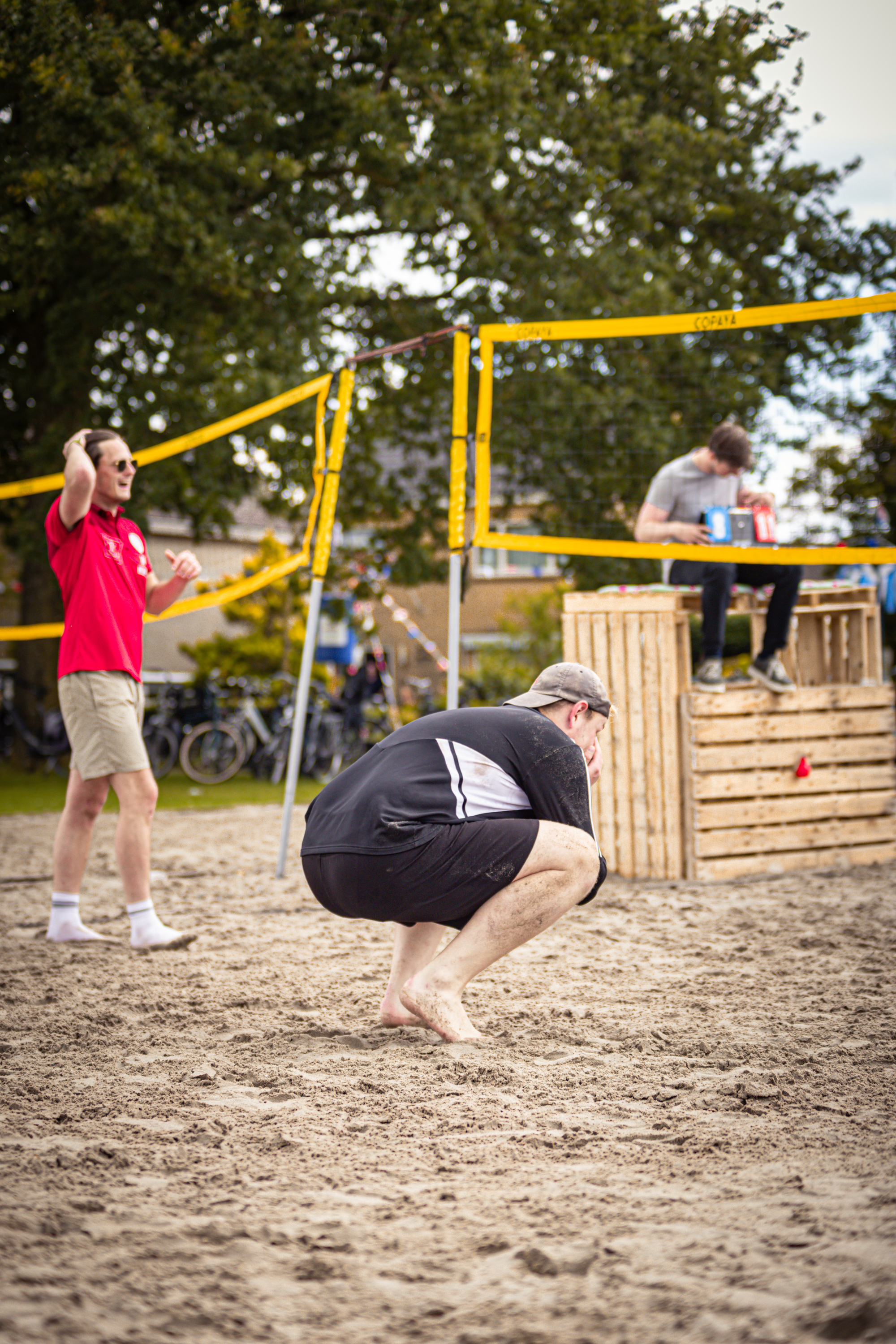 A man is playing volleyball on a beach in front of a man sitting on top of a pile of wooden planks.