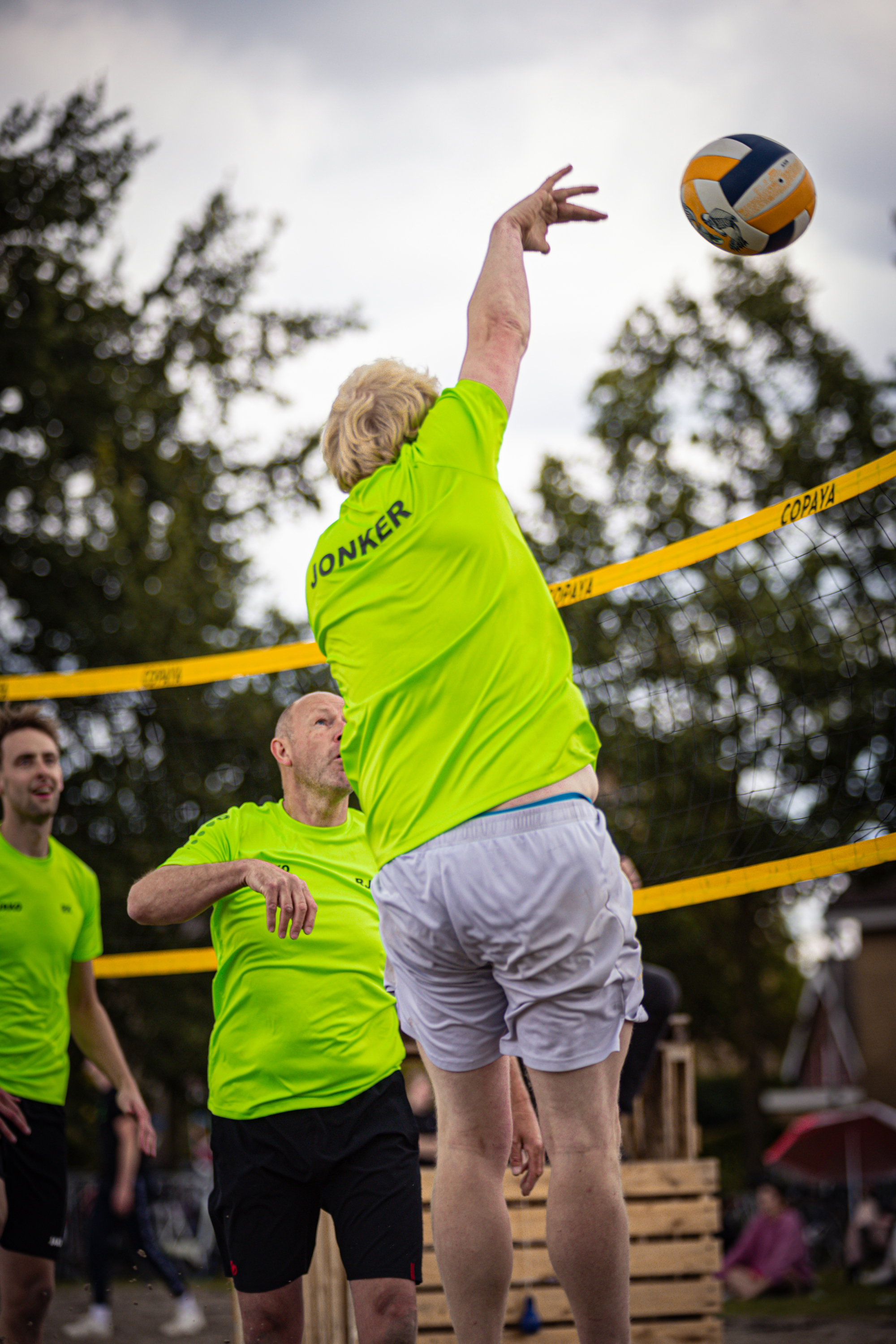 A man in a green shirt is jumping up to hit a yellow and blue beach volleyball.