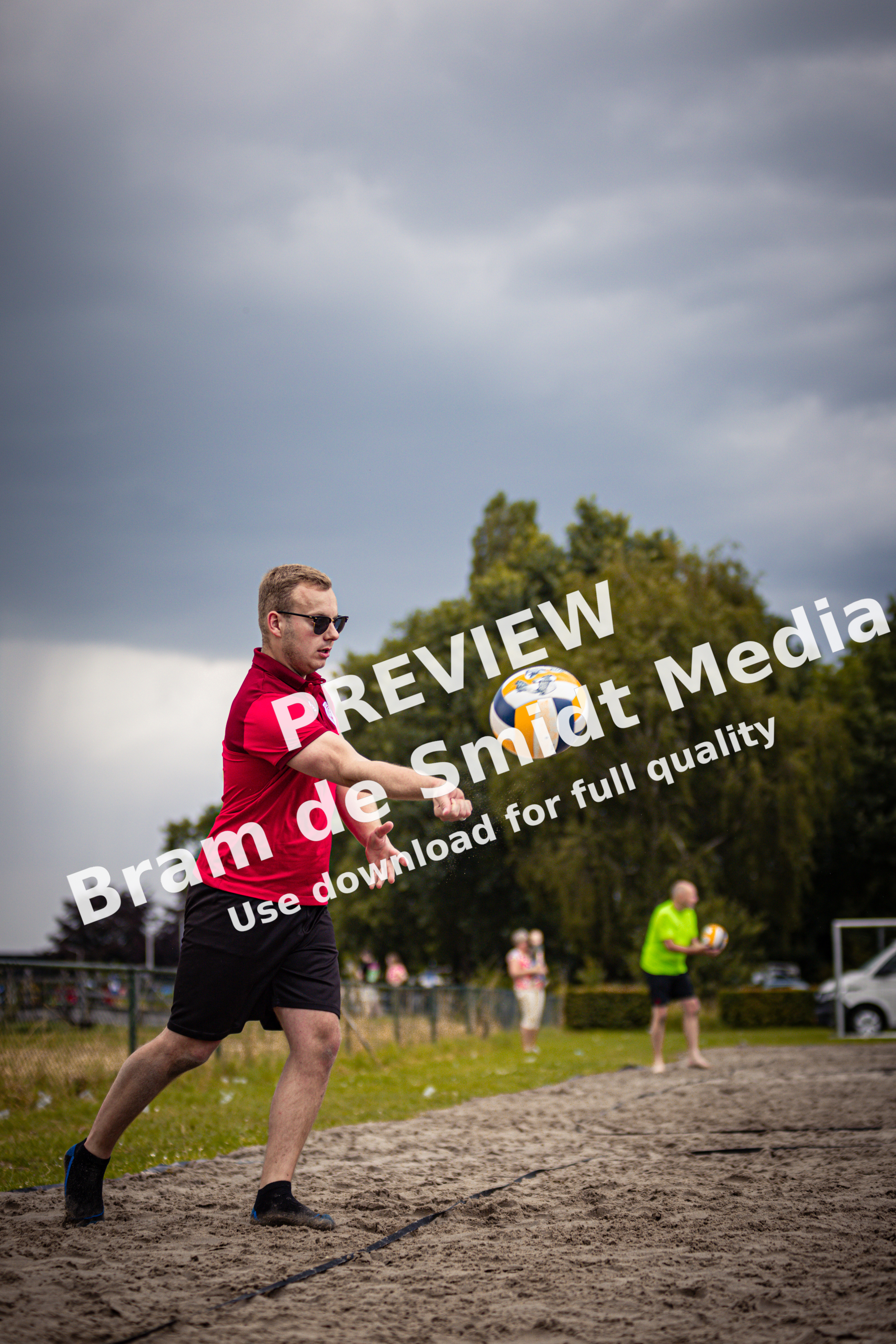 A man wearing sunglasses and a red shirt is playing beach volleyball with a ball on the court.