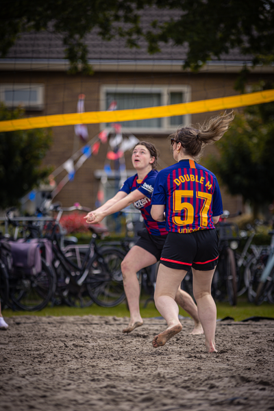 Two women are playing beach volleyball in a competition, the player with the blue jersey has the number 57 on it.