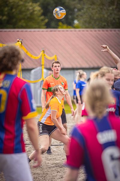 A group of people playing volleyball in the sand, one wearing an orange shirt and another a yellow one.