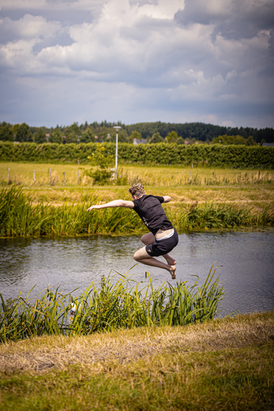 A man in a black shirt and shorts is playing volleyball on the grass near a pond.