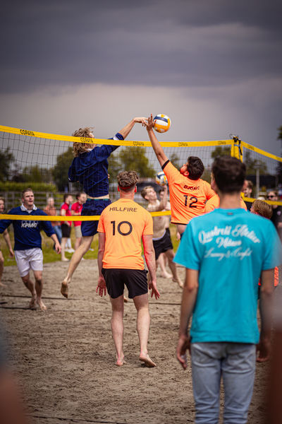 A group of young men playing beach volleyball, with one man wearing a blue shirt that says "Driestar Sport".