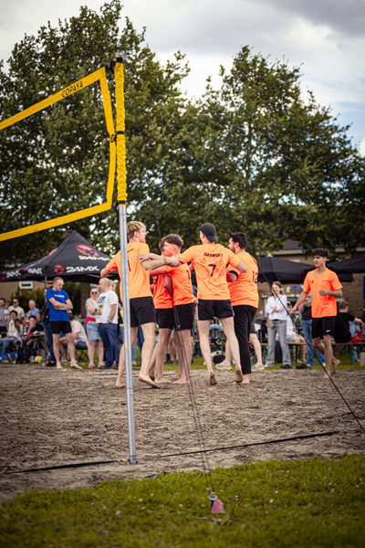 Five men in orange shirts holding hands and playing volleyball on a beach.