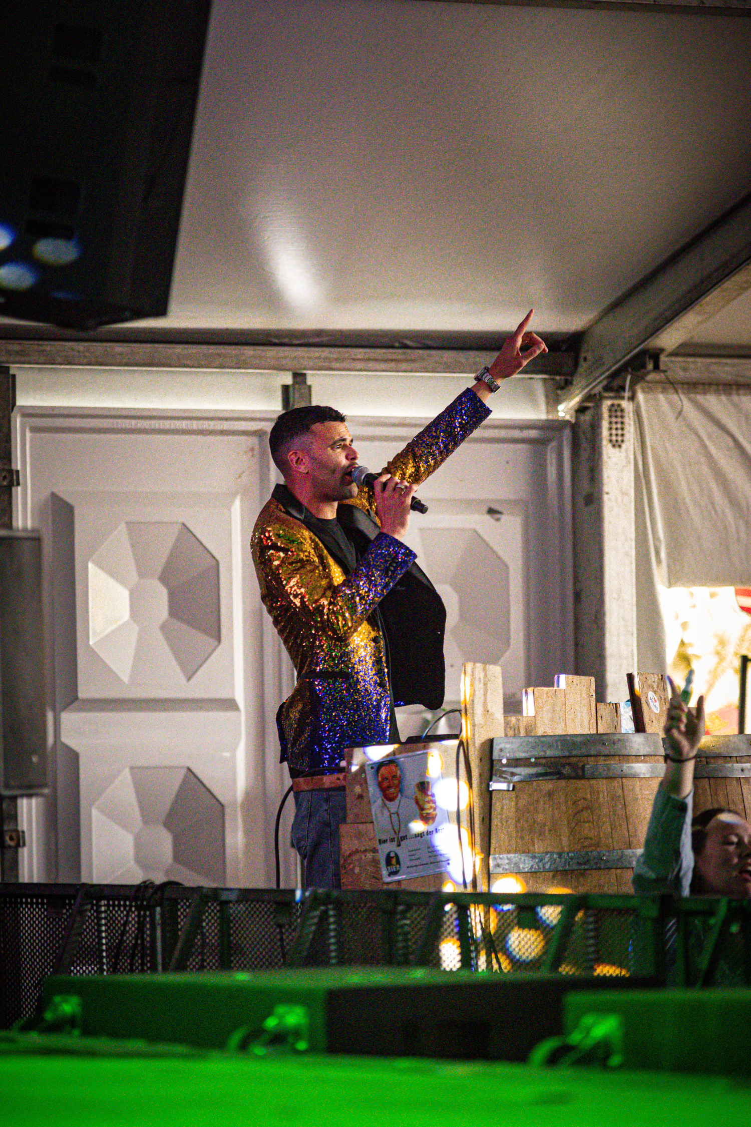 A man is on stage wearing a sequin jacket and has his arm raised.