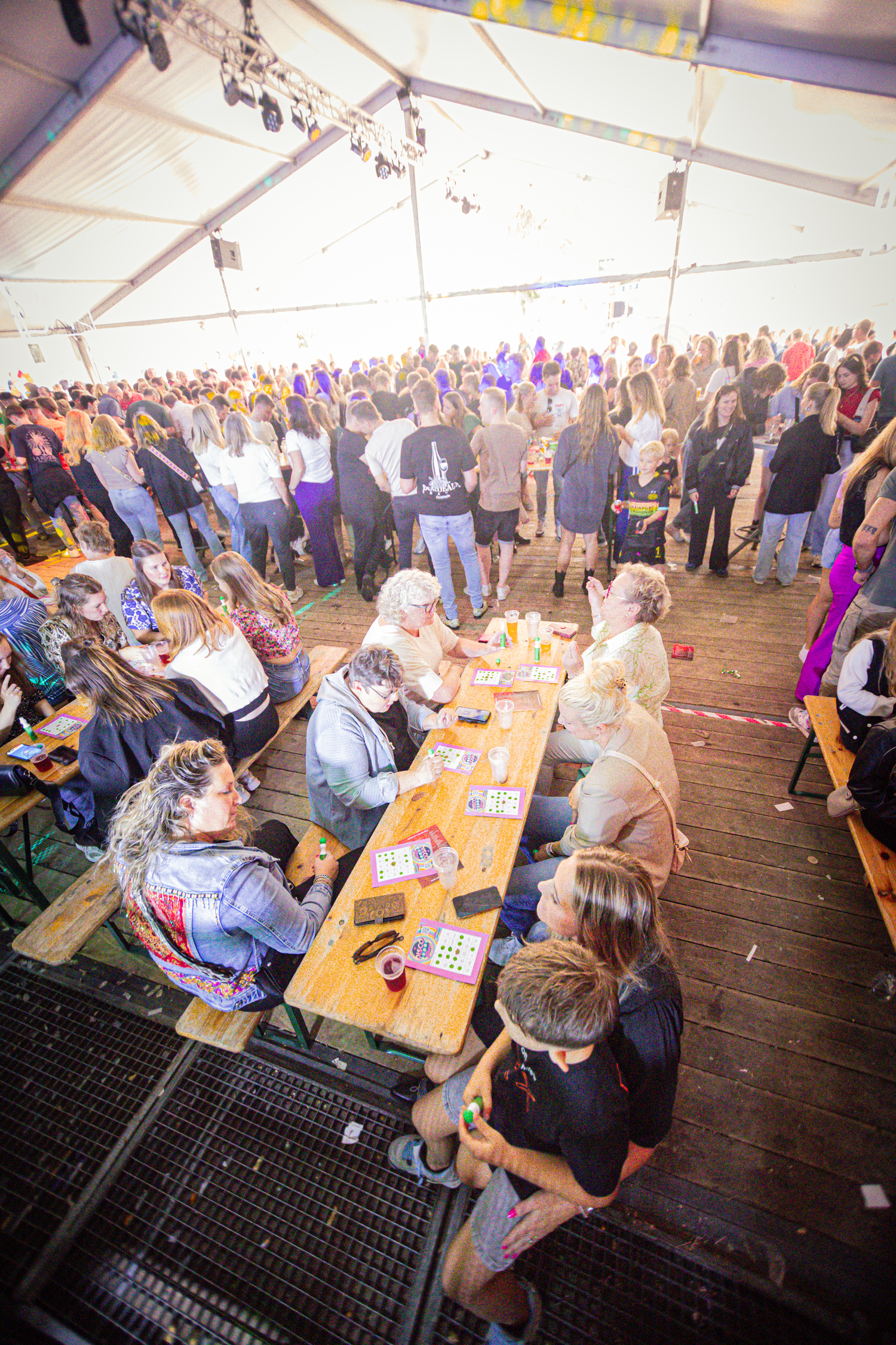 People at a festival are sitting around tables and chairs, some of them holding drinks.