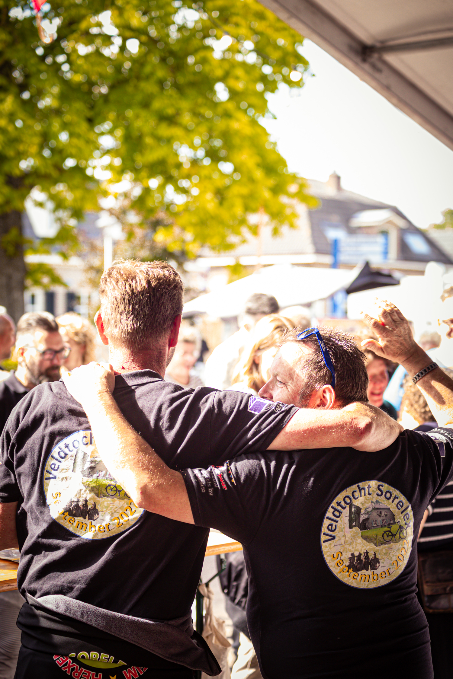 Two men are in a friendly embrace at a festival, one with a yellow logo on his shirt that says "Boerhaar".