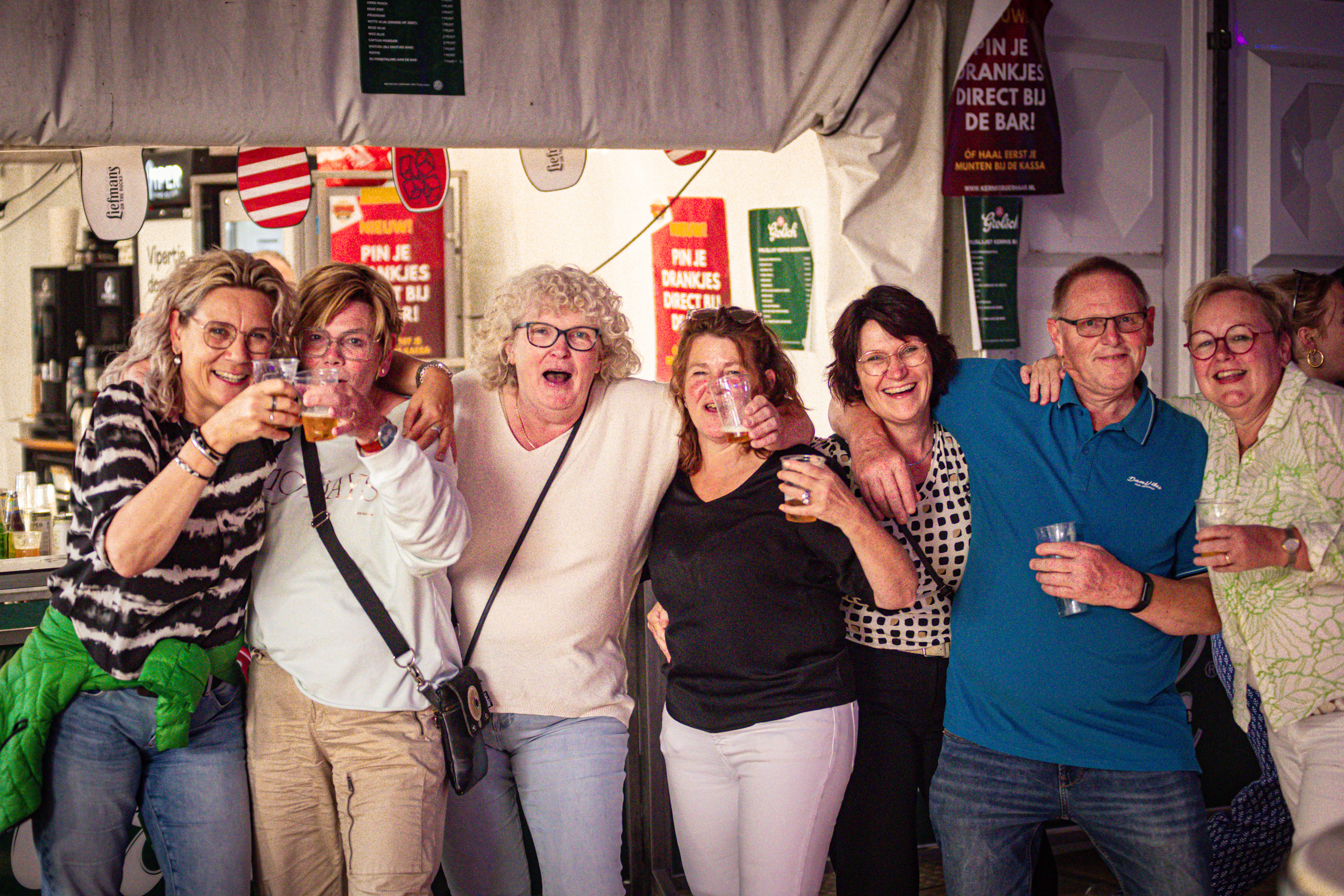 A group of people are posing for a photo with beer in their hands, one woman is wearing a shirt saying "Kermis Boerhaar".