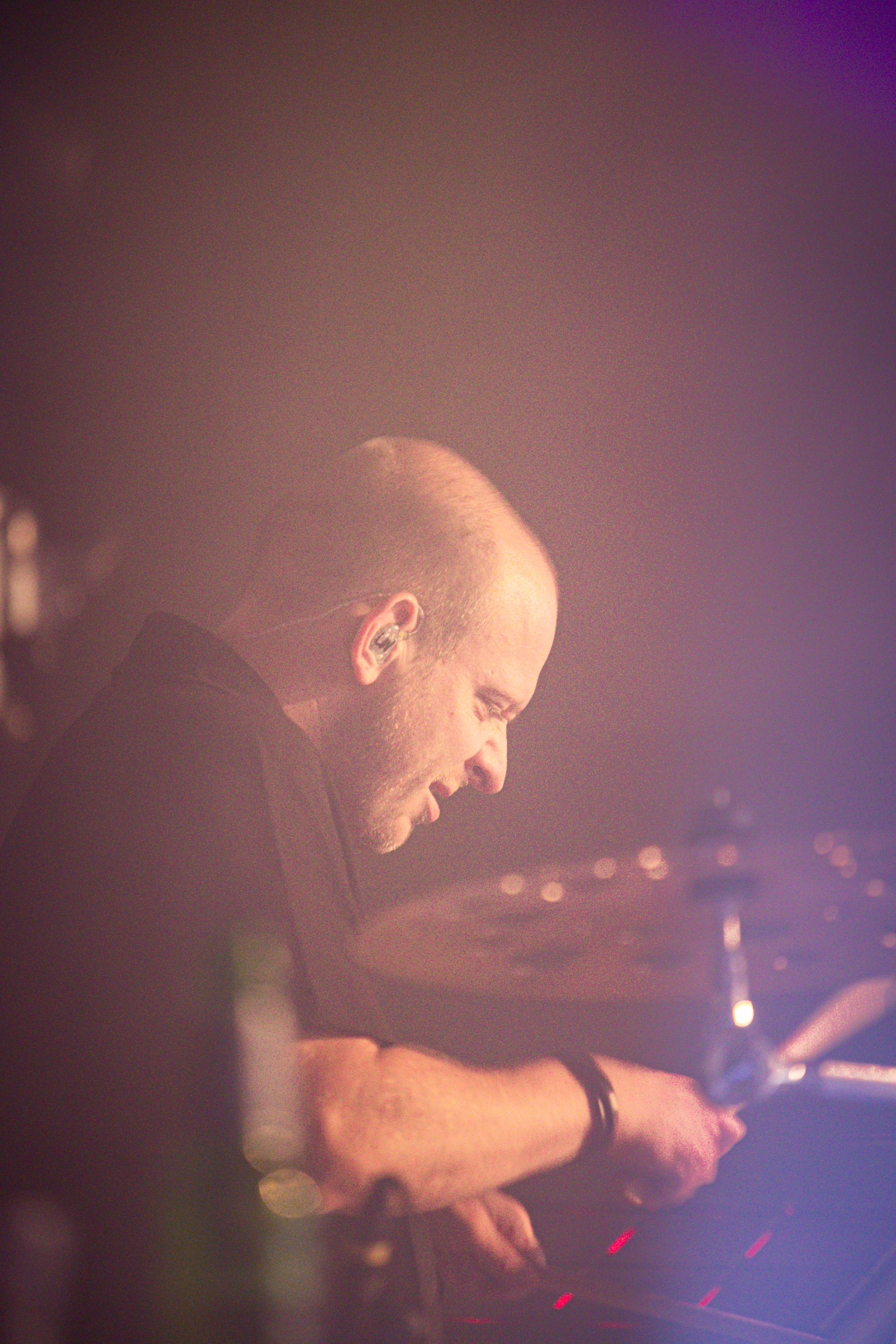 A man is sitting with a drum, holding a stick. The background is black and blurry.