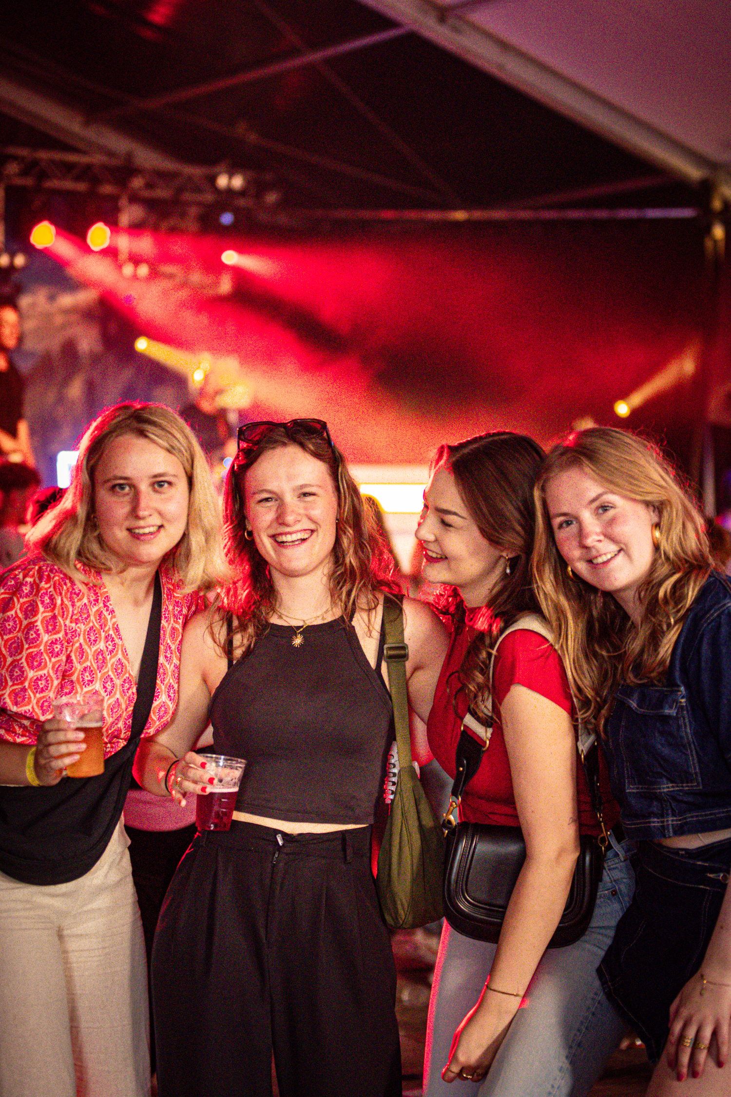 Four women posing for a photo together wearing casual clothing. The woman on the far left is holding a drink in her hand.