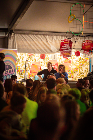 A large crowd of people gathered in front of a stage with a man on a microphone and a woman sitting at a table.