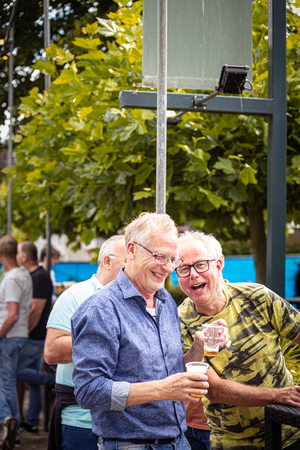 A group of people are standing outside under a sign, one man is wearing a blue shirt and the other is in a yellow shirt.