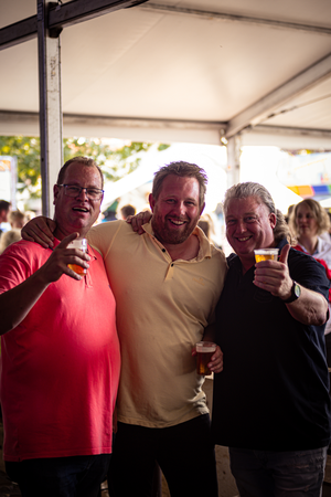 Three men posing at a festival, holding drinks and smiling.