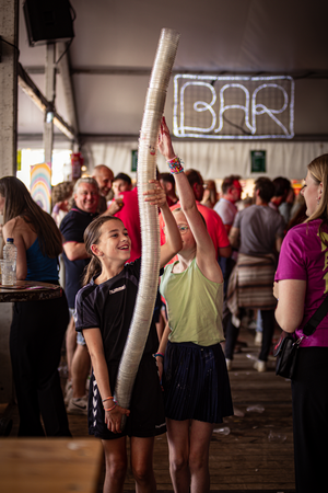 A young girl holding a stack of cups on her head, standing in front of a group of people at an outdoor event.