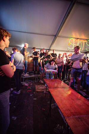 A group of people in a tent at an event called Kermis Boerhaar, with an orange table and a man with no shirt on.