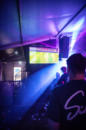 A man stands under a tent with a TV screen and speakers behind him. The words "Middag & avond" are visible on the screen.