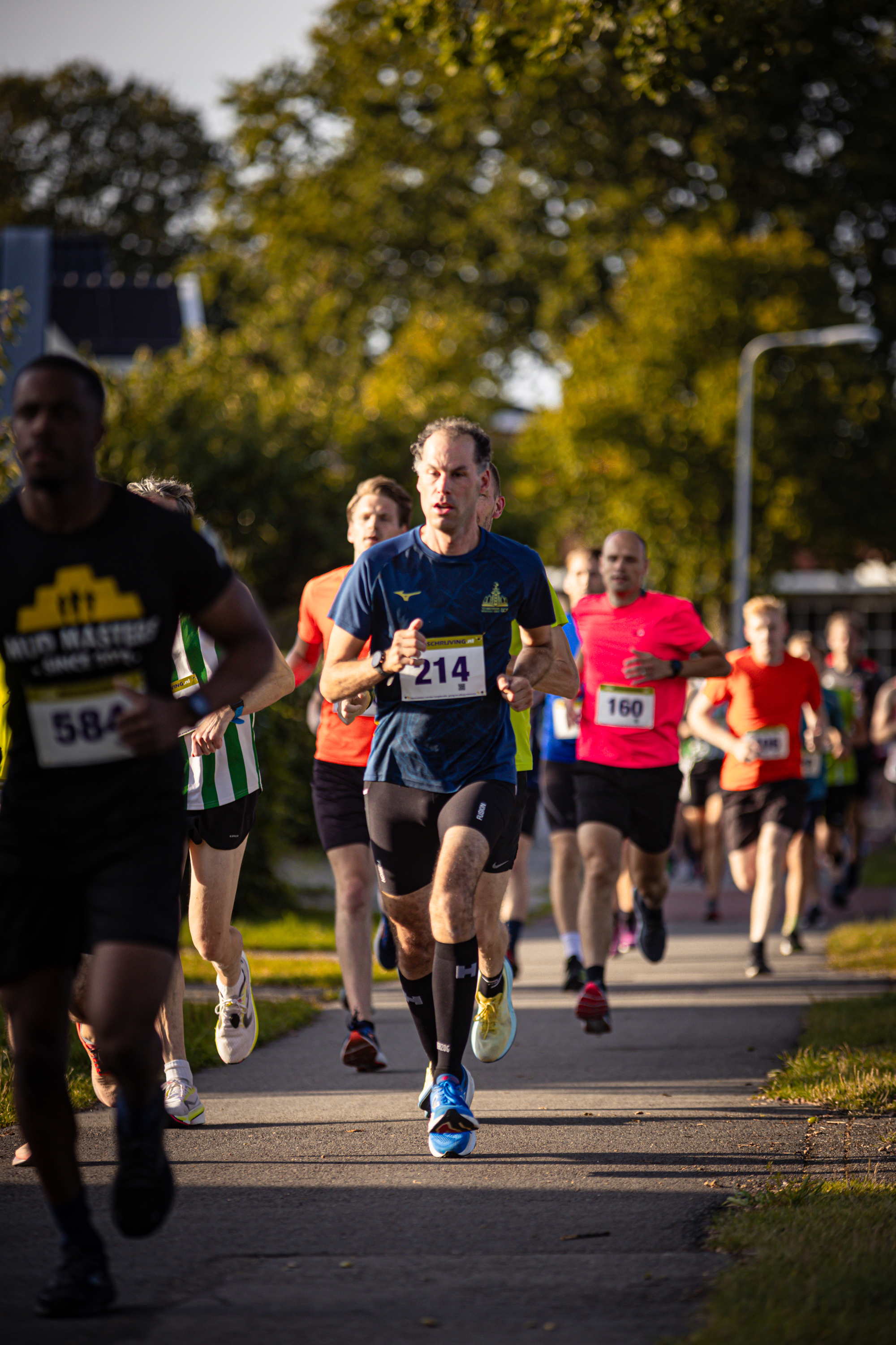 A group of people are running on a sidewalk, some of them wearing bibs with numbers such as 56 and 246.