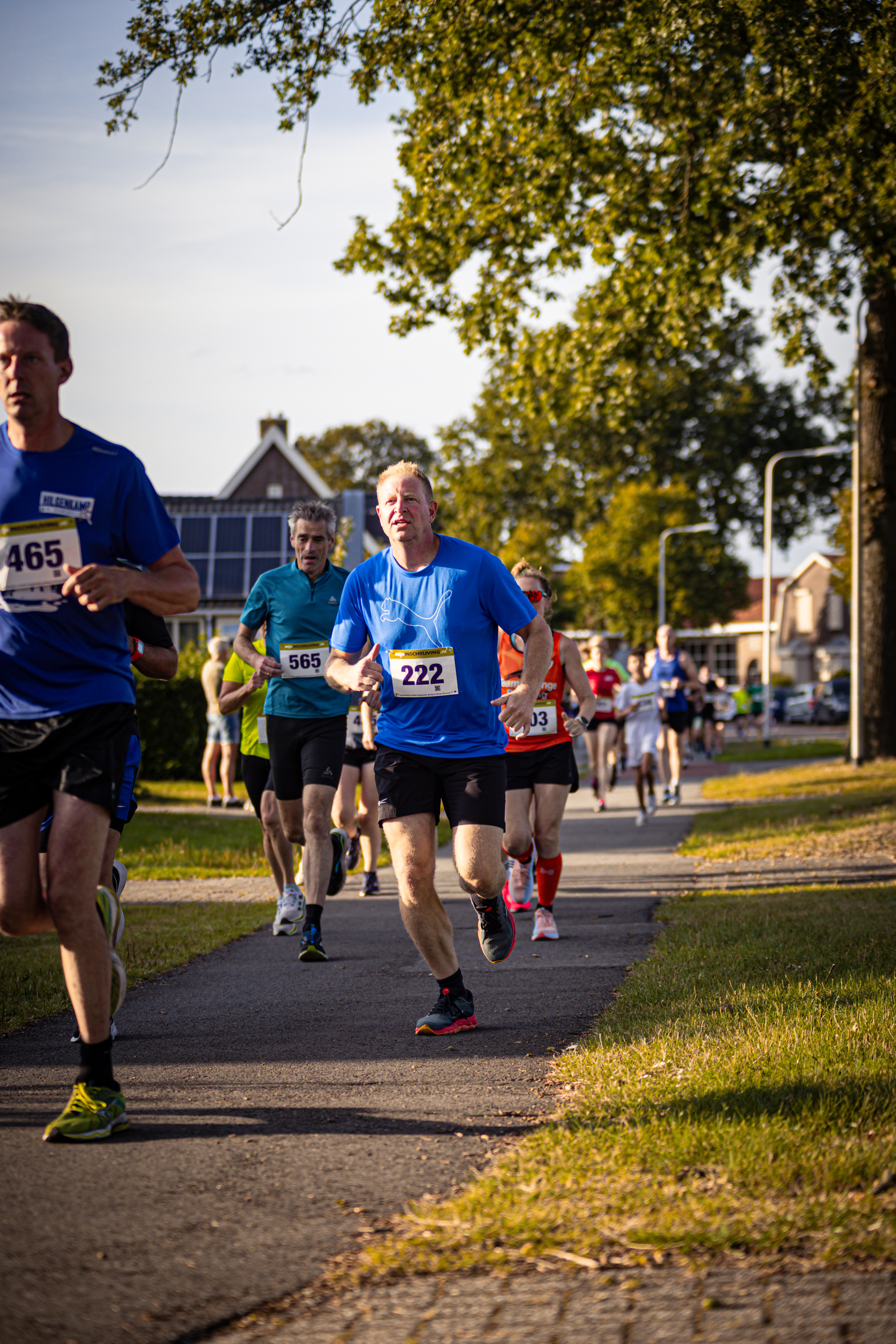 A man wearing a 225 number bib is running with other runners on a path near trees.