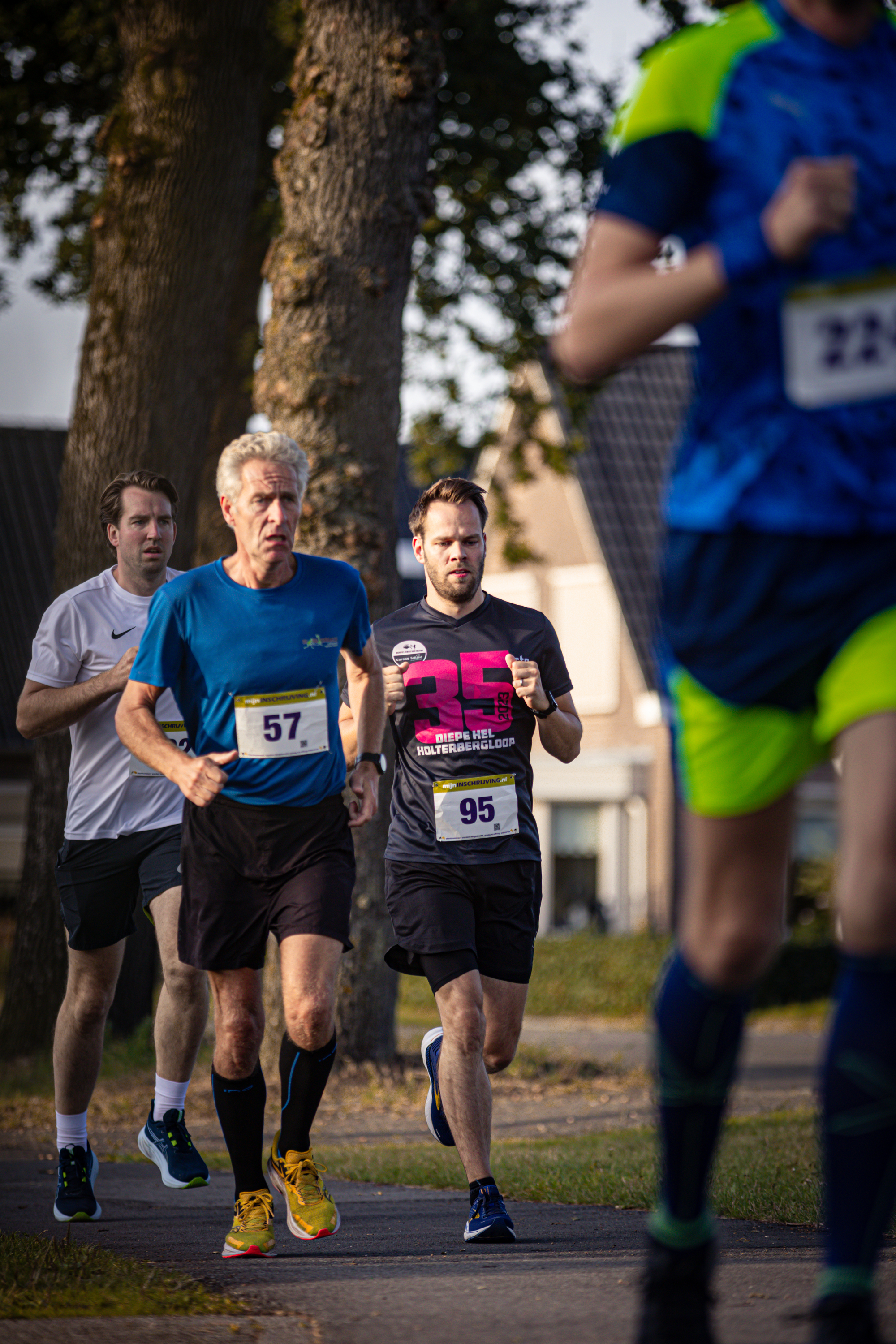 A man with a blue shirt and the number 57 on it runs behind two other runners.