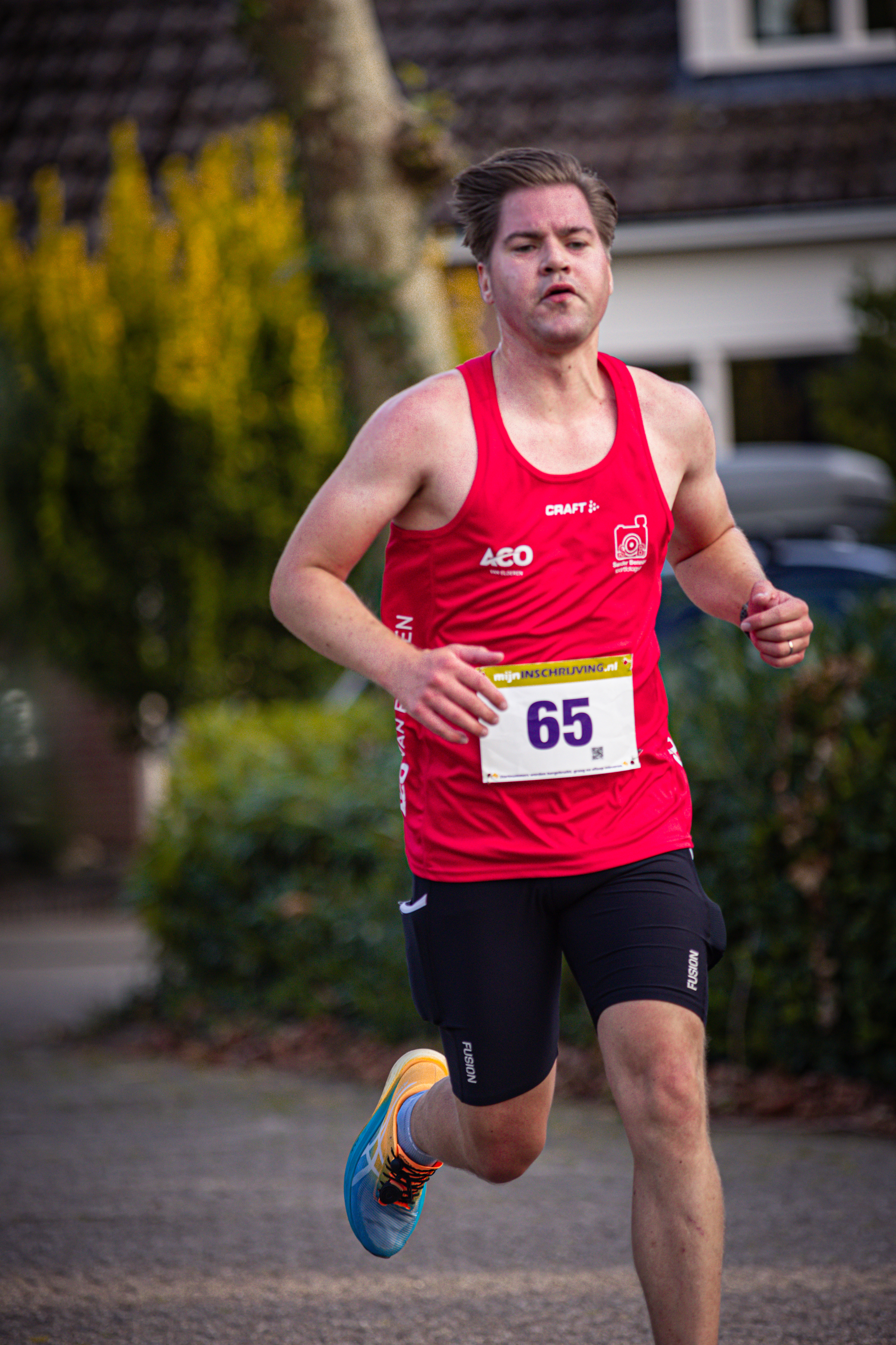 A male runner in a red tank top and blue shorts, running with the number 65 on his bib.